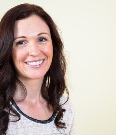 A woman with long, wavy brown hair smiles at the camera. She is wearing a light-colored shirt and stands against the plain, light beige Upper Walls of a Fremont studio.