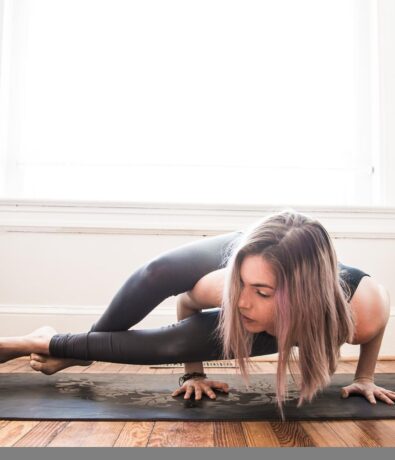 A woman with light brown hair practices a challenging arm balance yoga pose on a mat indoors, her body parallel to the floor and supported by her hands, legs extended to one side. Bright natural light fills the room.