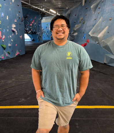 A man wearing glasses, a blue t-shirt, and beige shorts stands smiling in an indoor rock climbing gym with blue climbing walls and colorful holds, enjoying the perks of his summer membership.