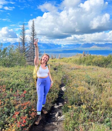 A woman in a white top and blue leggings stands on a mountain trail after climbing, smiling and raising one arm in a peace sign. She is surrounded by colorful wildflowers, trees, and distant mountains under a partly cloudy sky.