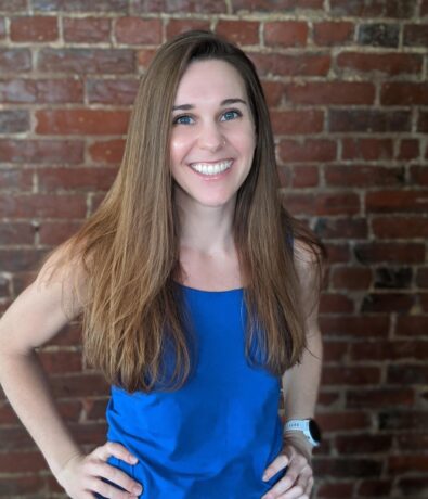 A woman with long brown hair, wearing a sleeveless blue top and a white smart watch, smiles confidently while standing in front of a brick wall, hands on her hips—ready for her next climbing adventure.
