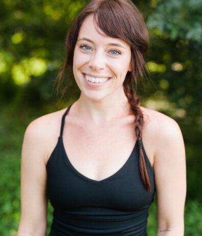 A woman with long brown hair in a side braid smiles at the camera. Wearing a black tank top, she stands outdoors amid greenery after a day of climbing.