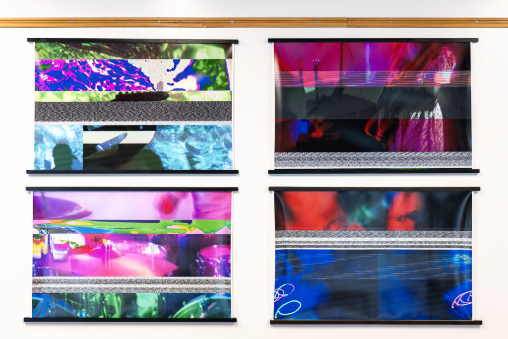 Four abstract digital artworks from a DC Arts community arts program fill the white wall with vibrant purples, blues, reds, and greens. Each piece features distorted shapes and horizontal glitch-like patterns arranged in two neat rows.