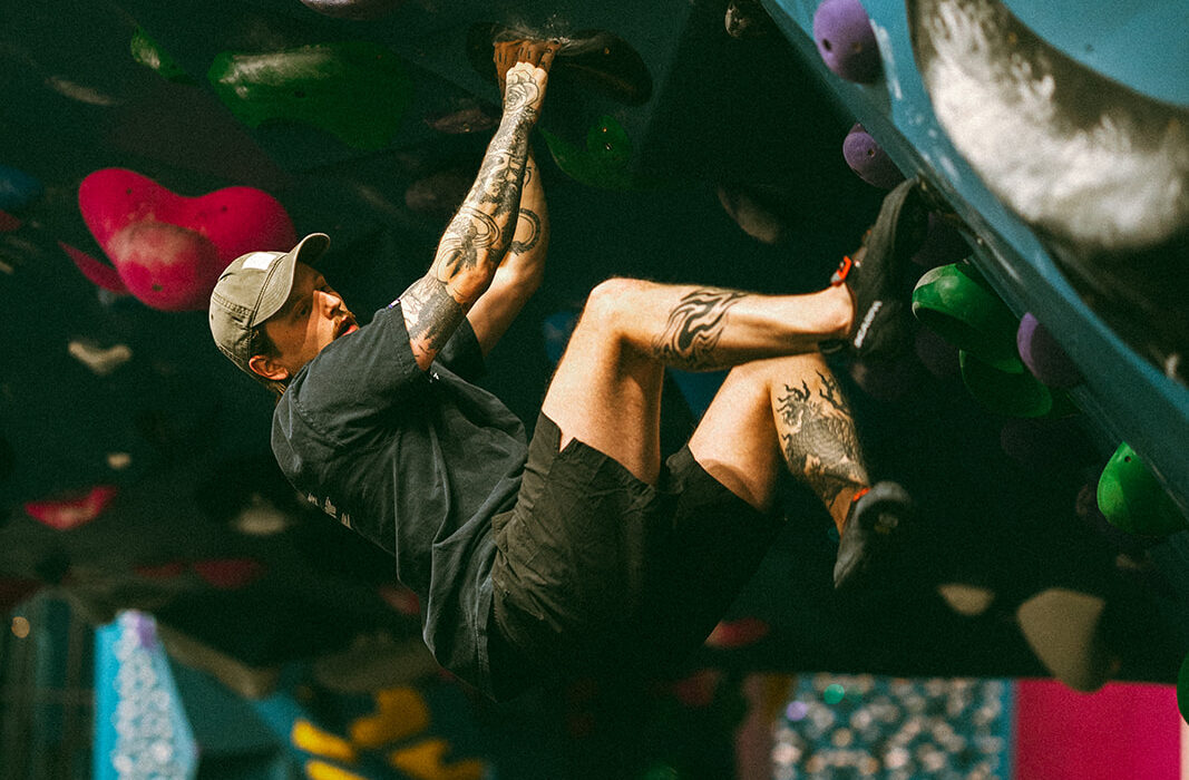 A person with tattoos and a cap climbs an indoor bouldering wall at a St Paul rock climbing gym, gripping holds on an overhanging blue surface. Their legs are bent as they hang close to the wall, focused on the next move.