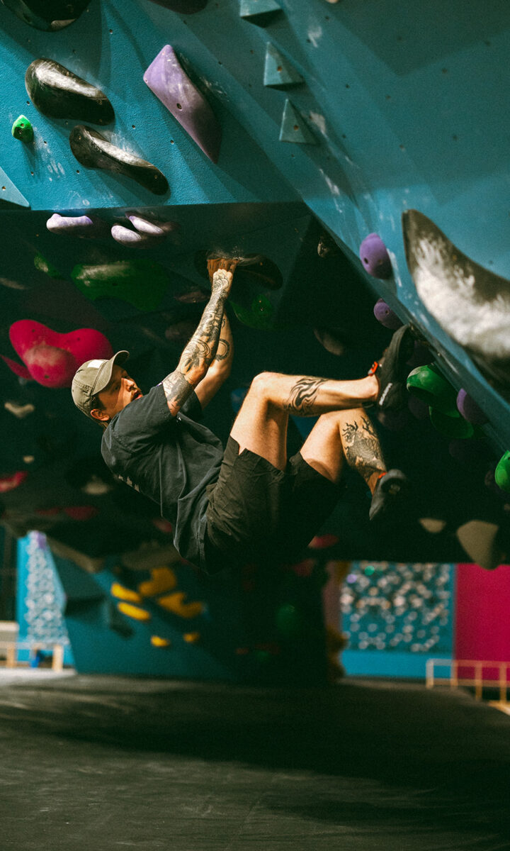A person with tattoos and a cap climbs an indoor bouldering wall at a St Paul rock climbing gym, gripping holds on an overhanging blue surface. Their legs are bent as they hang close to the wall, focused on the next move.