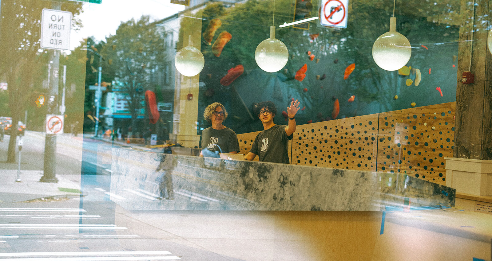 Two people stand behind a counter inside a modern building, smiling and waving. Large windows reflect a city street with trees and signs, creating a layered visual effect—perhaps welcoming or assisting with membership termination inquiries.
