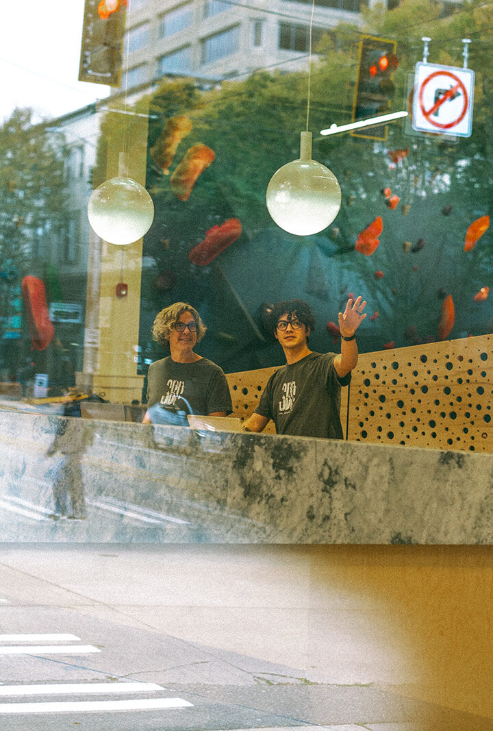 Two people stand behind a counter inside a modern building, smiling and waving. Large windows reflect a city street with trees and signs, creating a layered visual effect—perhaps welcoming or assisting with membership termination inquiries.