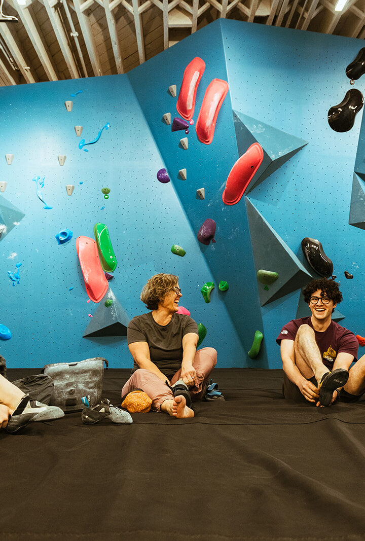 Three people sit on the floor of a climbing gym in Seattles University District, smiling as they put on their climbing shoes, surrounded by colorful climbing holds and vibrant walls in the background.