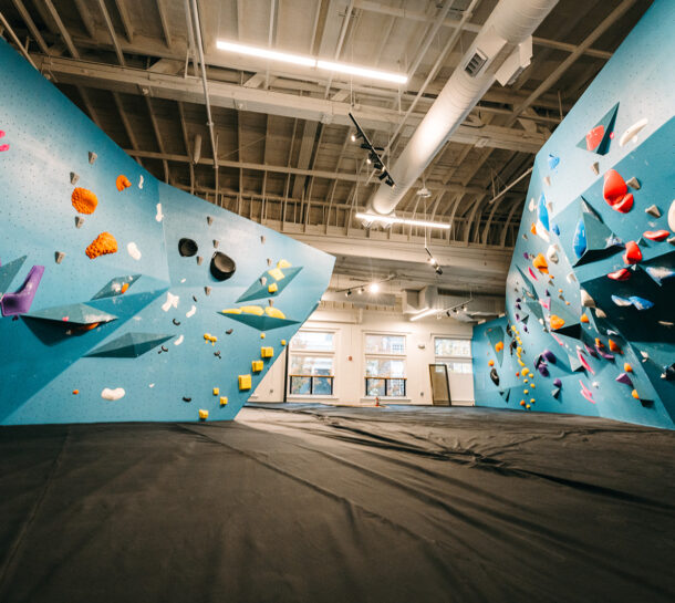 Indoor rock climbing gym in the U District featuring blue climbing walls, colorful holds, and large padded flooring. Enjoy a modern, open design with bright lights—visit during our Founding Opening Promo! No people are present in the scene.