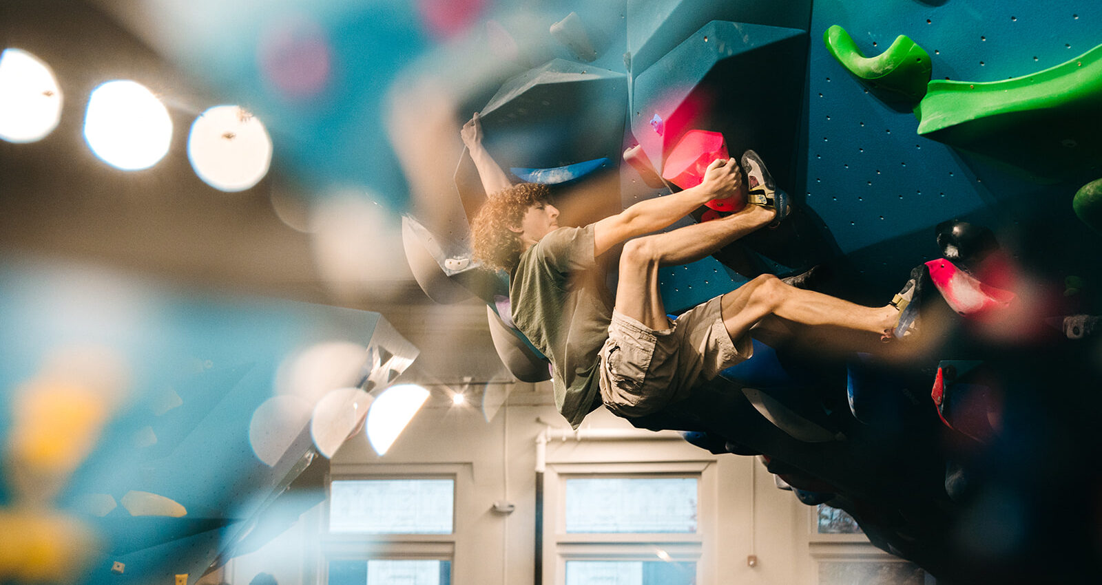 A person with curly hair climbs an indoor bouldering wall at a climbing gym in the University District, Seattle, gripping colorful holds. The image features reflections and blurred foreground, with windows and lights in the background.