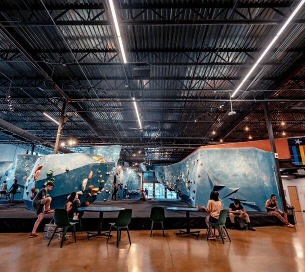 Indoor rock climbing gym with several people climbing on blue bouldering walls and others sitting at green chairs and tables. Overhead lights illuminate the Austin Climbing Gym, and a pride flag is visible on the wall to the right.