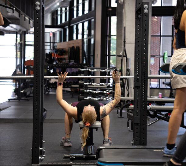 A person with tattoos lies on a bench under a barbell in a gym, preparing to lift weights. Another stands nearby, watching. The gym has climbing racks, benches, and large windows in the background.