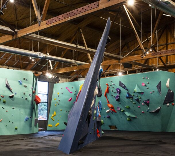 A modern indoor climbing gym in Fremont featuring light blue and green Upper Walls, colorful holds, and a large overhanging wall in the center beneath wooden ceiling beams and exposed ducts.