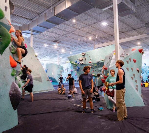 People are indoor rock climbing and bouldering in a spacious gym, with groups talking, preparing, and climbing on colorful climbing walls under bright lights. The atmosphere appears active and social.