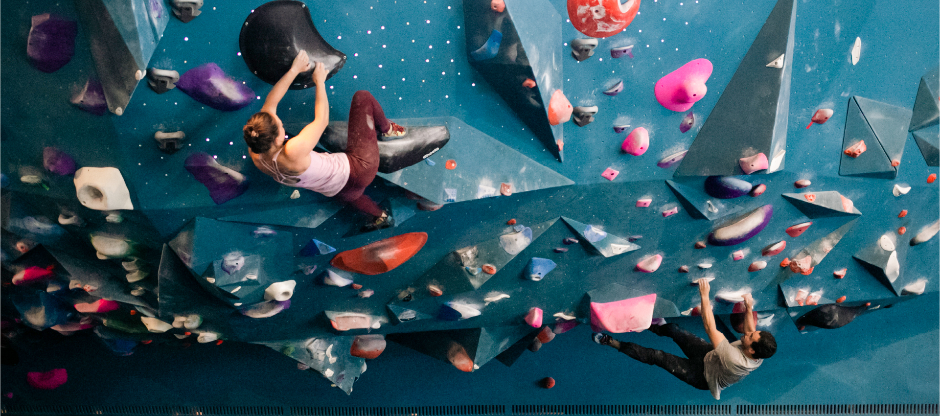 Two people climb on an indoor bouldering wall with colorful holds and geometric features. One climber is near the top left, the other lower right. Both appear focused, scaling the challenging blue wall.