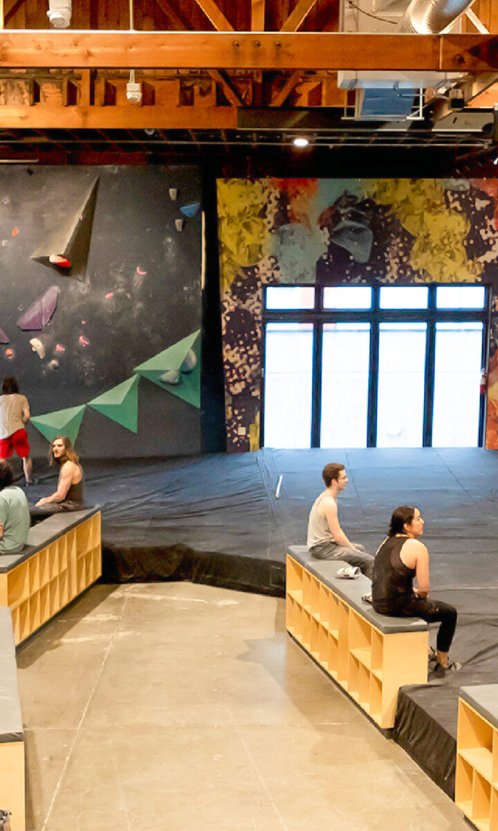 Indoor bouldering gym in Fremont with colorful climbing holds on angled Upper Walls, wooden ceiling beams, and a few people sitting or climbing. Natural light enters through large windows at the far end.