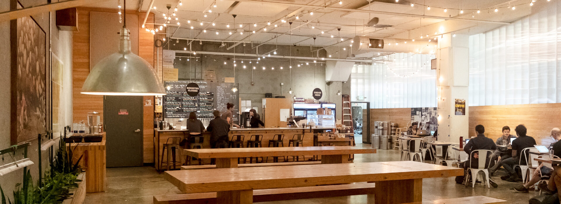 Spacious modern café in Fremont with wooden tables, string lights, and a long bar counter. Some people work on laptops; others stand at the counter. The Upper Walls add charm to the cozy, casual atmosphere.