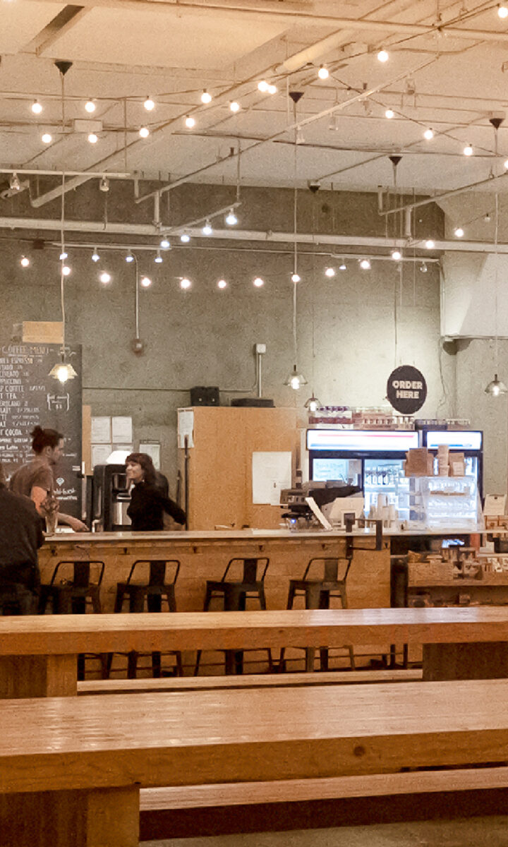 Spacious modern café in Fremont with wooden tables, string lights, and a long bar counter. Some people work on laptops; others stand at the counter. The Upper Walls add charm to the cozy, casual atmosphere.