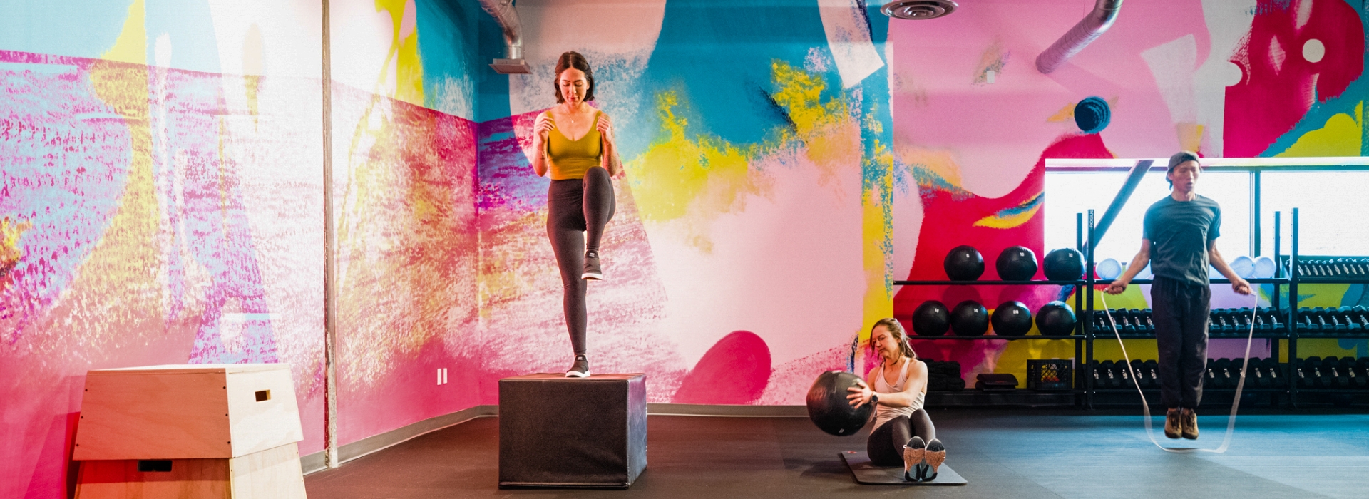 Three women exercise in a colorful gym: one steps onto a box, one sits and holds a medicine ball, and one jumps rope. Racks with weights and vibrant abstract wall art are in the background.