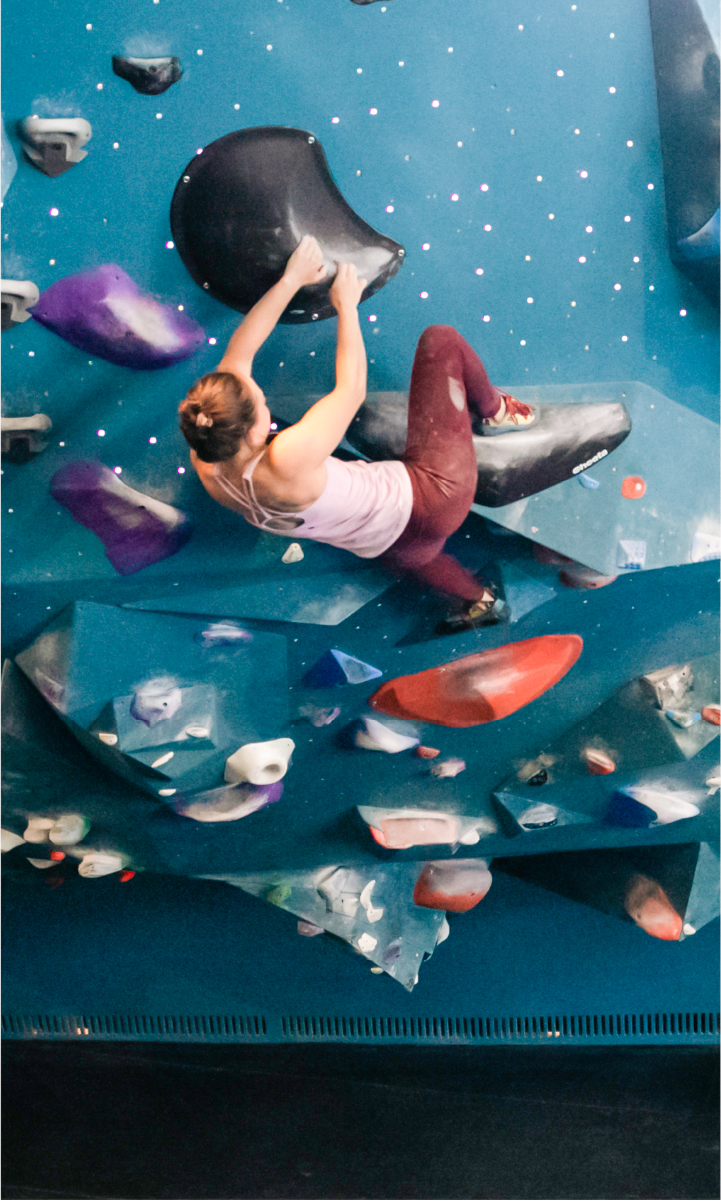 A woman wearing a white tank top and maroon leggings climbs a blue indoor bouldering wall, gripping a large black hold with both hands and balancing her feet on various colored holds.