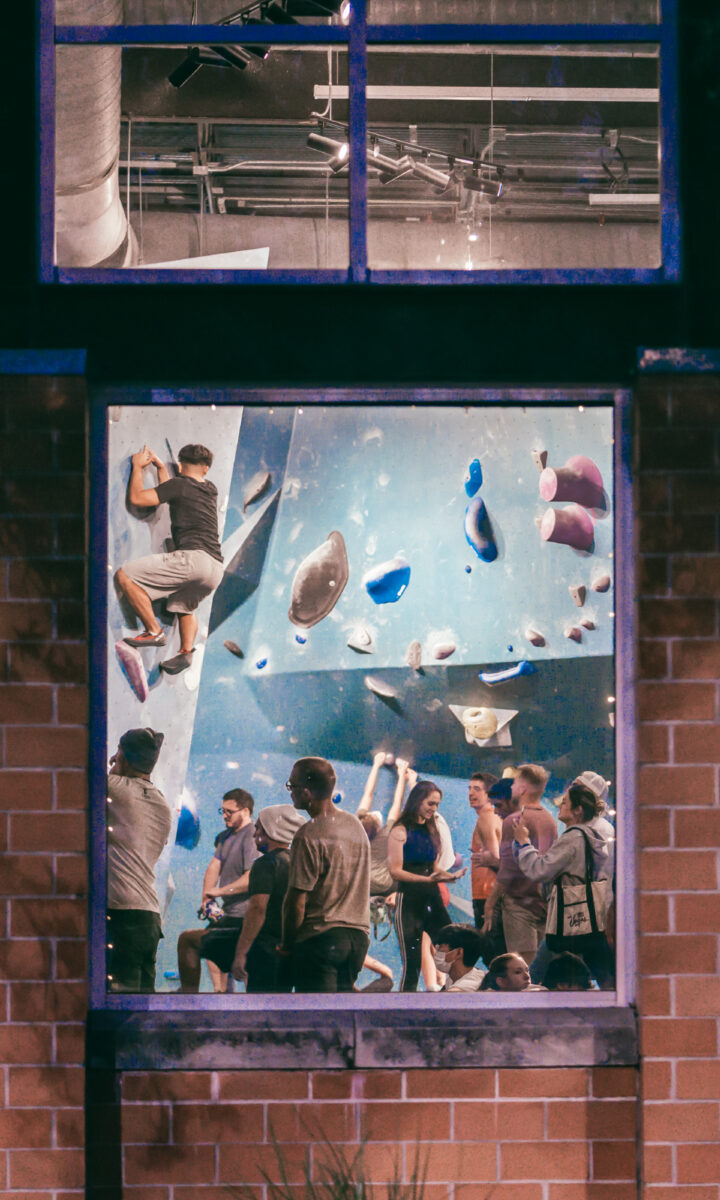 People socialize and watch as someone climbs an indoor bouldering wall, seen through a large window from outside a building at night. Various climbing holds are visible on the blue wall.