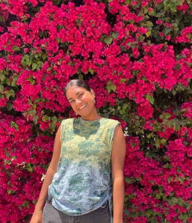 A woman smiles while standing in front of a lush wall of vibrant pink climbing bougainvillea flowers. She wears a sleeveless green and blue patterned top and dark pants, as bright sunlight illuminates the scene.