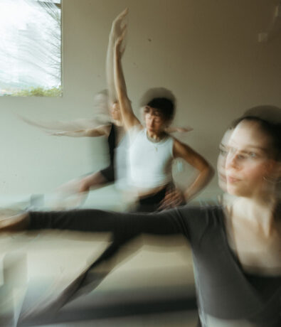 Three women practice ballet in a studio near the University District Seattle, their movements captured with a motion blur effect, creating a sense of graceful motion and fluidity. Soft, natural light filters through a window in the background.