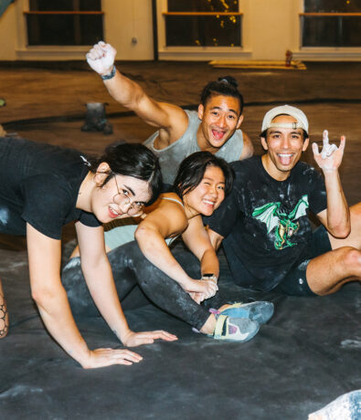 Four young adults covered in chalk smile and pose playfully on a climbing gym floor during a Climb for Community event. Two crouch in front, two sit behind—one flashing a rock-on gesture—with climbing holds and gym windows in the background.