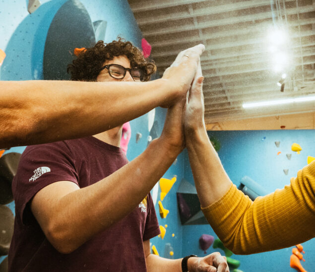 Three people give a group high-five in front of a colorful indoor climbing wall during a Climb for Community event. The focus is on their hands, with one person wearing glasses and curly hair visible in the background.