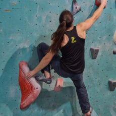 A person with long brown hair, wearing a black sleeveless shirt and climbing shoes, is scaling an indoor bouldering wall, reaching up with one arm toward a red hold.