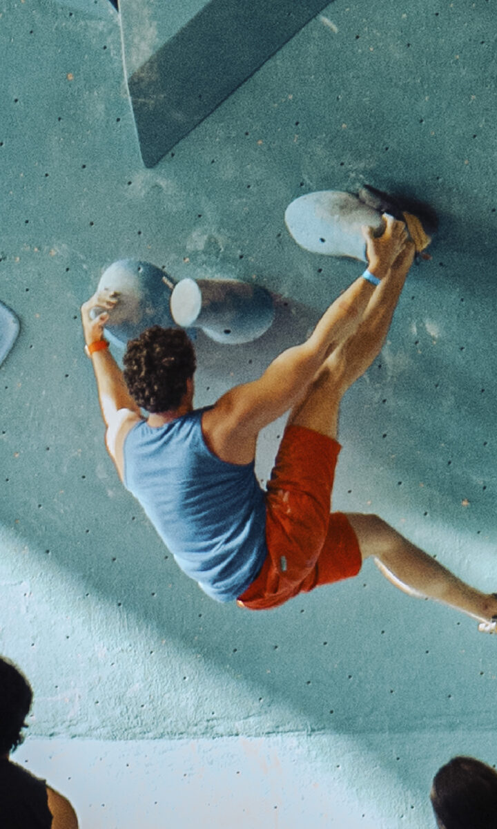 A man in a blue tank top and orange shorts climbs an indoor bouldering wall with colorful holds, as a group—some chatting about the new Membership Freeze option—watches from below, some standing and others sitting.