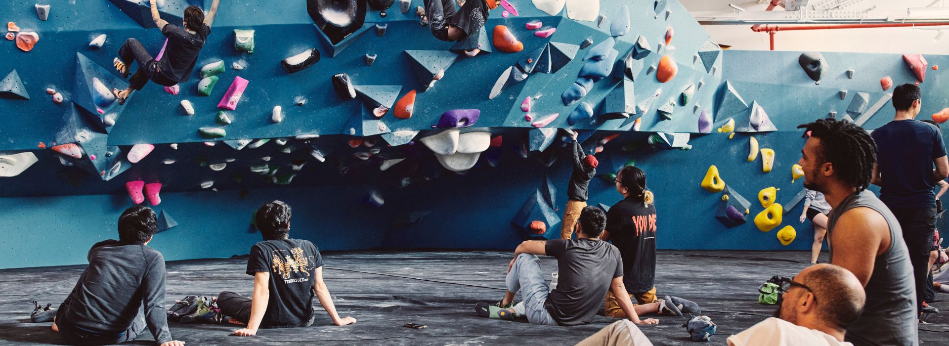 People gather on padded flooring to watch climbers tackle colorful holds at a lively indoor bouldering gym. Groups chat and cheer, capturing the vibrant energy typical of Brooklyn events and Arcteryx events in town.