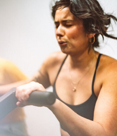 A woman in a black sports bra exercises intensely on a rowing machine, gripping the handle tightly and showing strong focus and determination on her face.