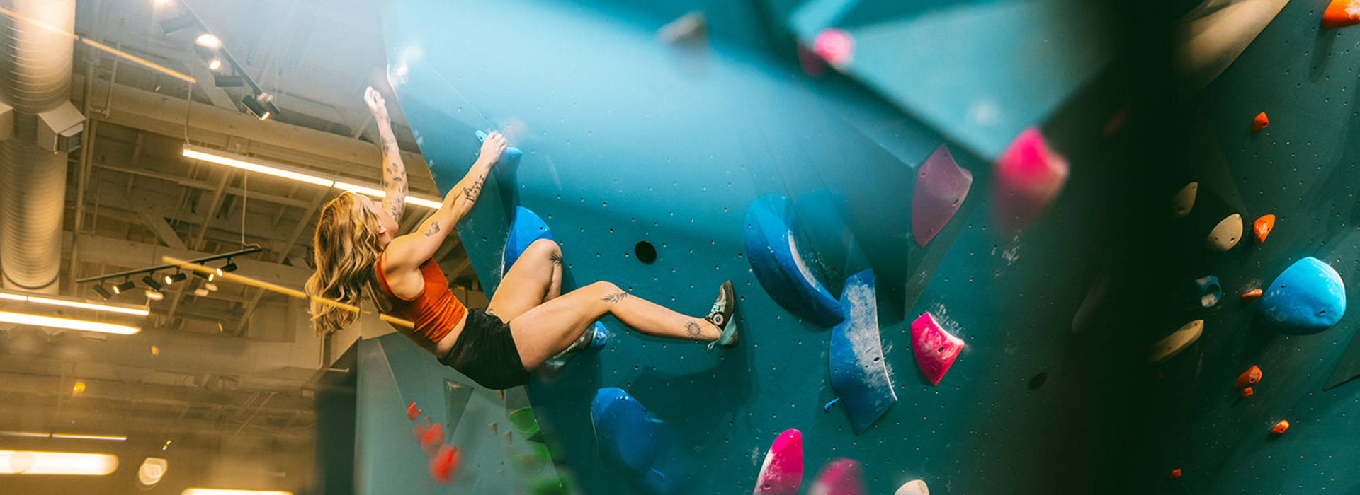 A person with tattoos climbs an indoor bouldering wall, gripping colorful holds. They wear a red tank top, black shorts, and climbing shoes, their hair loose in a brightly lit gym—perfect for making the most of a summer membership.