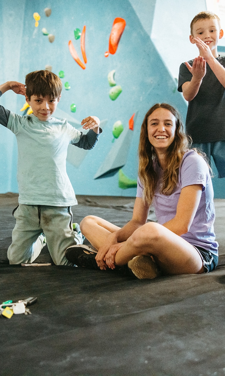Two boys pose and a young woman smiles while sitting on the floor of an indoor climbing gym during Parents Night Out, with colorful climbing holds visible on the wall behind them.