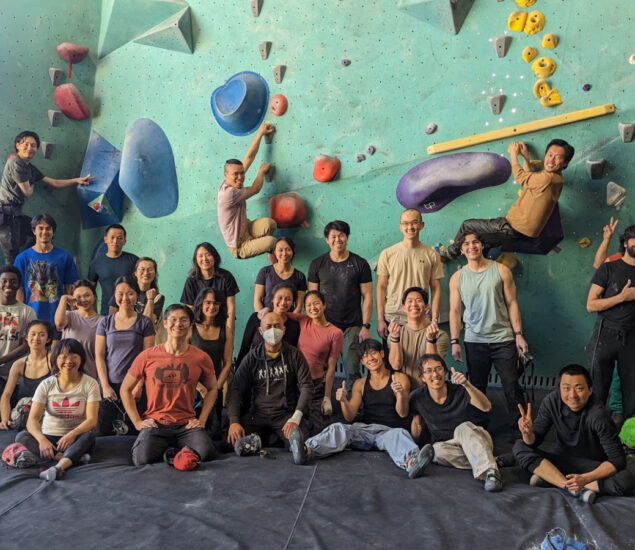 A large group of people from local Community Clubs pose and smile together in front of an indoor rock climbing wall in Minneapolis. Some stand, while others sit or kneel on the padded floor, and a few climb holds on the wall in the background.