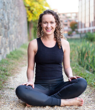 A woman with curly hair sits cross-legged on a gravel path outdoors, smiling at the camera. Dressed in a black tank top and leggings, she looks ready for a day of climbing, with greenery and buildings in the background.