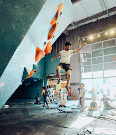 A person in mid-air after jumping off an indoor climbing wall with orange holds, while several people watch and others sit or stand on padded flooring in a bright, spacious gym.
