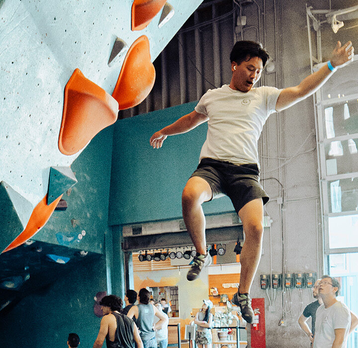 A man in a white t-shirt and shorts is falling or jumping off an indoor climbing wall with orange holds, mid-air, in a busy climbing gym with people watching—perfect for those enjoying their summer membership.