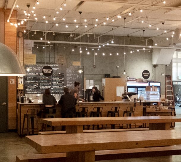 A cozy bar with string lights overhead, several people seated at the counter, and wooden tables in the foreground. The menu is written on blackboards behind the bar, and a Register Closed sign is visible.