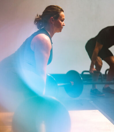 A woman in athletic wear is lifting a barbell in a gym, with another person lifting weights in the background. Soft, colorful light flares partially obscure the scene.