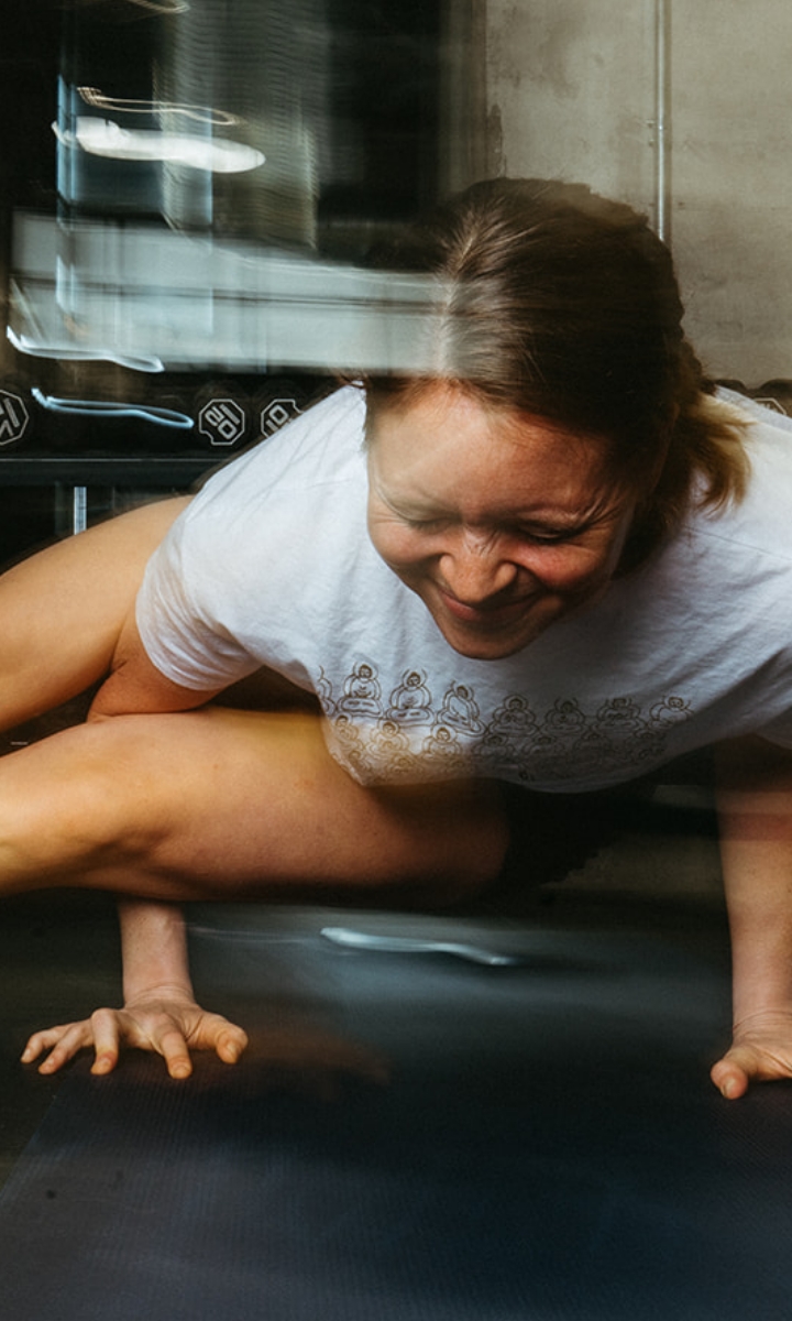 A person in a white t-shirt is performing a challenging yoga pose indoors, balancing on their hands with one leg over their arm and a focused, smiling expression—perfect for showcasing our Membership benefits and Pricing options. The image has a dynamic, blurred effect.