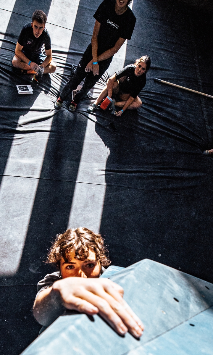A child climbs a wall in a sunlit gym while three other children sit on the floor below, watching and smiling. The scene is dynamic with shadows and light streaming across the floor, capturing the excitement of membership activities.