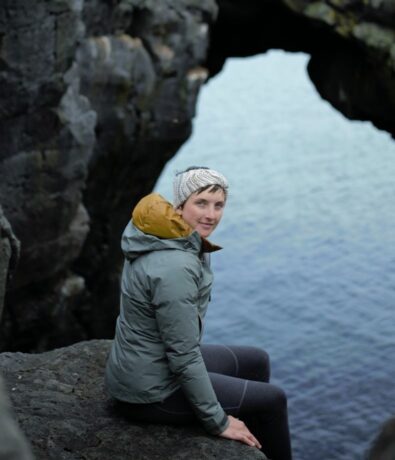A woman in a gray jacket and headband sits on a rocky ledge near the edge of a cliff, with blue water and a large rock arch visible in the background.