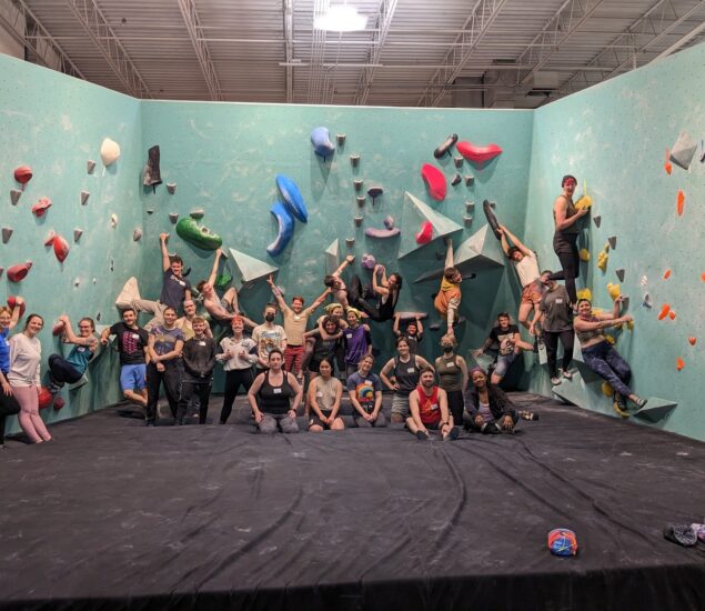 A large group of people pose and smile in front of an indoor bouldering wall in Minneapolis; some stand on the ground while others climb or sit on holds, showcasing colorful climbing routes and a strong sense of community clubs.