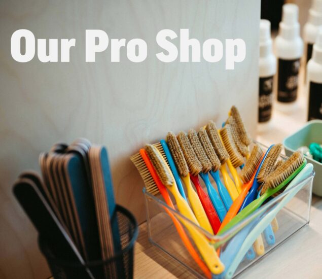 A clear container holds assorted cleaning brushes with colorful handles, perfect for any retail space. Nearby are emery boards in a holder and bottles on a shelf. Large white text reads “Our Pro Shop” on a light wooden background.