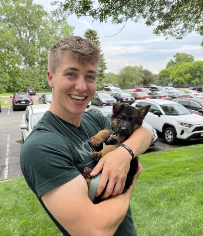 A smiling young man stands on a grassy area in front of parked cars, holding a small black and brown puppy in his arms. Trees and a partly cloudy sky are visible in the background.