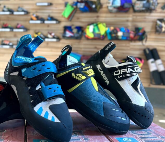 Three pairs of colorful rock climbing shoes are displayed side by side on a table in this retail setting, with a blurred background showing various MBP climbing gear on wooden shelves.