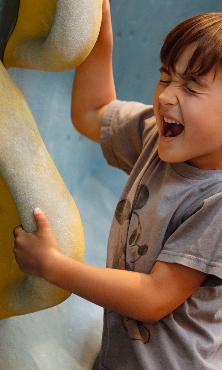 A young boy with brown hair and a gray Mickey Mouse shirt smiles with his eyes closed while gripping large, yellow climbing holds on an indoor climbing wall in Brooklyn during the school holidays.