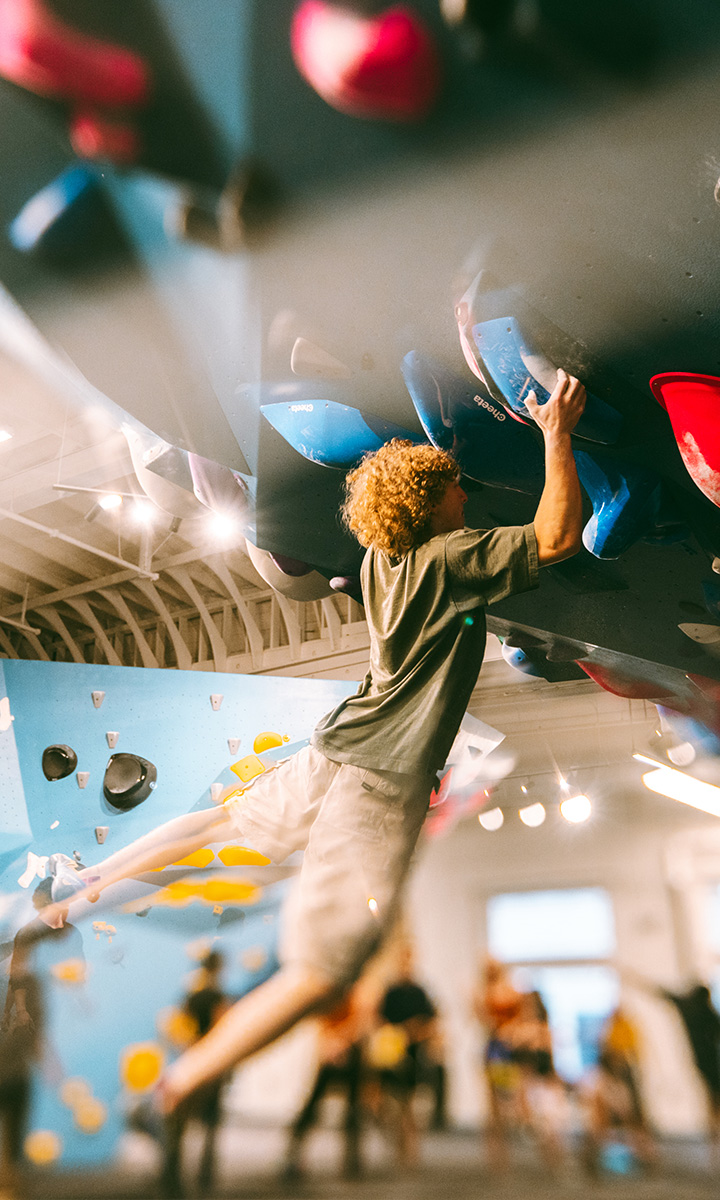 A person with curly blond hair climbs an indoor bouldering gym wall at Climbing Tempe, gripping blue handholds with one hand while stretching their body mid-motion. The background shows other climbers and colorful holds.